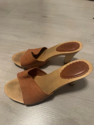 Bruna träklackade sandaler - Säljer ett par bruna sandaler med träklack och öppen tå. Ovandelen är i brunt skinn med dekorativa sömmar och nitar på sidorna. Klacken är hög och i trä, vilket ger en retro känsla. Perfekta för dig som gillar en boho-vibe och vill ha något unikt till sommaren.