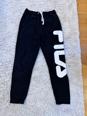 Svarta mjukisbyxor från FILA - Svarta mjukisbyxor från FILA med stor vit logga längs ena benet och detaljer i rött och vitt på baksidan. Byxorna har resår i midjan med vit snörning och fickor på sidorna samt en bakficka. Perfekta för chill eller träning.