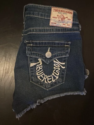True Religion denimshorts Joey Cut-Off - Säljer ett par mörkblå denimshorts från True Religion, modell Joey Cut-Off. Shortsen är lågmidjade med råa, fransiga benslut och har stora bakfickor med broderad logga. Klassiska silversfärgade knappar och dragkedja framtill. Inga defekter, vid frågor är det bara att skriva💕