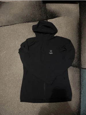 Haglöfs vindjacka  - Snygg svart hoodie från Haglöfs med dragkedja framtill och diskret logga på bröstet. Hoodien har en klassisk huva och två sidofickor. Tillverkad i mjukt material som passar perfekt för chill eller utomhusaktiviteter.