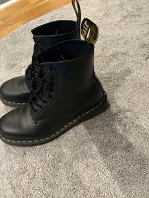 Svarta Dr. Martens kängor i skinn - Klassiska svarta Dr. Martens kängor i skinn med gul söm runt sulan och snörning framtill.  Storlek 39. Använda ca 2-3 ggr så väldigt bra skick 