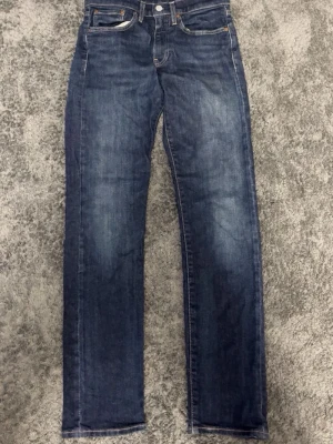 Levi's 501 blå raka jeans - Klassiska blå Levi's 501 jeans med rak passform och femficksdesign. Jeansen har en tidlös look med knappgylf och subtila slitningar på benen. Perfekta för dig som gillar en enkel och stilren denimstil.