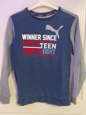 Blå och grå långärmad tröja Puma - Snygg långärmad tröja från Puma i blått med grå ärmar och rygg. Framsidan har tryck med texten 'Winner Since Nineteen Forty Eight' i vitt, rött och blått samt Puma-logga. Rund halsringning och mjukt material, perfekt för en sportig look.