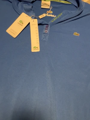 Blå pikétröja från Lacoste - Snygg blå pikétröja från Lacoste med klassisk krage och knappar framtill. Tröjan har det ikoniska krokodilmärket broderat på bröstet och en liten Lacoste-tag i sidan. Tillverkad i ett mjukt pikématerial som känns skönt mot huden. Det är storlek S. Pris kan diskuteras vid snabb affär. Först till kvarn!