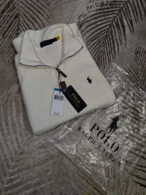 Vit halvzip från Polo Ralph Lauren - Klassisk vit halvzip-tröja från Polo Ralph Lauren med broderad marinblå logga på bröstet. Tröjan har hög krage, dragkedja med läderdetalj och är tillverkad i mjukt material som känns lyxigt. Perfekt för en clean och stilren look.