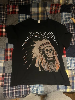 Svart Yeezus t-shirt med tryck - Svart t-shirt med Yeezus-tryck fram och bak, stor dödskalle med fjäderskrud på framsidan och turnéstäder på baksidan. Klassisk passform och rund hals. Perfekt för dig som gillar streetwear och statement-plagg. Storlek XL sitter mer som L