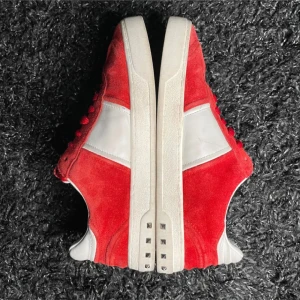 Röda och vita sneakers med nitar - Snygga sneakers i röd mocka och vitt skinn med coola silverfärgade nitar på sulan. Skorna har klassisk snörning och en chunky vit sula. Perfekta för dig som vill sticka ut med en unik detalj på fötterna.