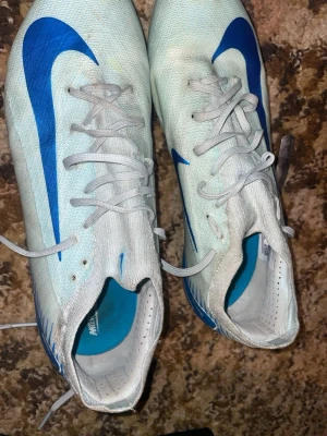 Nike vita och blå fotbollsskor - Säljer ett par vita Nike fotbollsskor med blå detaljer och snörning. Skorna har en låg profil och är tillverkade i syntetmaterial för lätt känsla på planen. Perfekta för dig som vill ha stilrena och snabba skor till matchen.