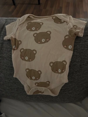 Beige body med björnmönster - Supersöt beige body med bruna björnhuvuden som mönster. Kortärmad och mjuk i materialet, perfekt för små barn. Tryckknappar i grenen för enkel påklädning.