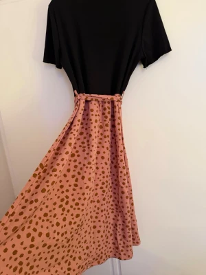 Svart och rosa prickig klänning SHEIN - Supersnygg klänning från SHEIN med svart ribbad överdel och korta ärmar. Kjoldelen är rosa med bruna prickar och har en knytrosett i midjan. Lång modell som ger en flowy känsla och passar perfekt till vår och sommar.