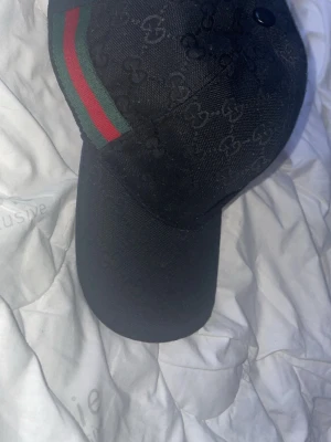 Svart Gucci keps med GG-mönster - Säljer en svart Gucci keps med klassiskt GG-mönster över hela kepsen och ikoniska gröna och röda ränder på sidan. Justerbar rem bak i skinn och insida med Gucci-etikett. Perfekt accessoar för dig som vill ha något stilrent och lyxigt.