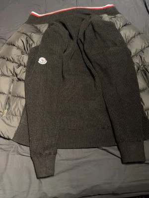 Svart bomberjacka från Moncler - Snygg svart bomberjacka från Moncler med quiltad framsida och stickade ärmar. Jackan har dragkedja framtill, Moncler-logga på bröstet och ribbade muddar. Perfekt för dig som vill ha en stilren och exklusiv look.