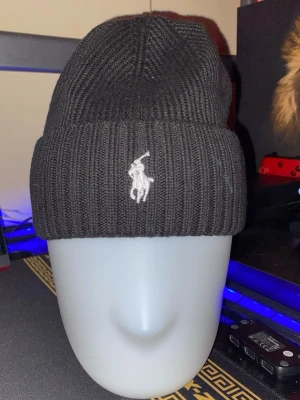 Svart ribbstickad mössa från Ralph Lauren - Svart ribbstickad mössa från Polo Ralph Lauren med klassisk broderad logga i vitt framtill. Tjockt uppvik och mjukt material som håller värmen. Perfekt accessoar för kalla dagar och snygg till streetwear-stil.