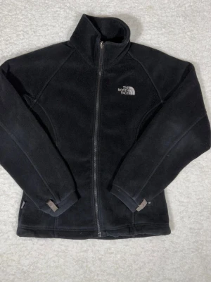 Svart fleecejacka från The North Face - Mysig svart fleecejacka från The North Face med dragkedja framtill och hög krage. Jackan har broderad logga på bröstet och praktiska muddar vid ärmsluten. Perfekt för lager-på-lager och kyliga dagar.