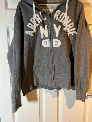 Abercrombie & Fitch hoodie - Säljer en grå zip-hoodie från Abercrombie & Fitch som är mycket trendiga och snygga inga defekter! Storlek XL men passar mig som är 183 och väger ca 65kg så hade sagt runt M egentligen. Vid frågor är det bara och höra av dig!