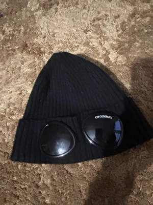 Svart mössa med goggles - Säljer en svart ribbstickad mössa med ikoniska inbyggda goggles framtill. Mössan har en tight passform och är tillverkad i mjukt material som håller värmen. Perfekt för dig som vill sticka ut med en streetwear-inspirerad look.