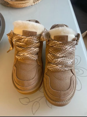 Beige sneakers från UGG med snörning - Säljer ett par beige sneakers från UGG med grova snören i beige och vitt mönster. Skorna har en rund tå, platt sula och är fodrade med mjukt vitt foder. Ovandelen är i textil och mocka för en schysst look.