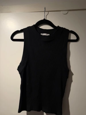 Svart ribbad ärmlös topp - Snygg svart ribbad topp med hög hals och ärmlös design. Perfekt att styla med jeans eller kjol för en trendig look. Materialet är stretchigt och följsamt för en bekväm passform.