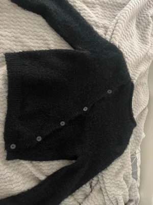 Svart stickad kofta från NA-KD - Mysig svart stickad kofta från NA-KD med asymmetrisk knäppning framtill. Koftan har långa ärmar, rund halsringning och är gjord i ett fluffigt material som ger en trendig look. Perfekt för lager på lager och enkel att styla till olika outfits.