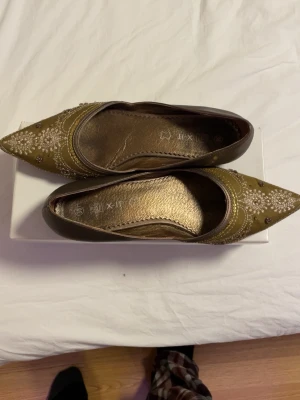 Guldiga ballerinaskor med spetsig tå - Snygga ballerinaskor i guld med spetsig tå och dekorativa broderier samt pärldetaljer. Skorna har en platt sula och är tillverkade i syntetmaterial med glittriga inslag. Perfekta för dig som vill sticka ut med en unik och elegant stil.