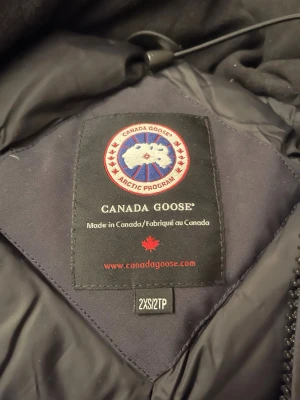 Svart dunjacka från Canada Goose - Snygg svart dunjacka från Canada Goose med klassisk patch på ärmen och stor huva med pälsdetalj. Jackan har dragkedja och knappar framtill, perfekt för kalla vinterdagar. Ikonisk design och riktigt varm.