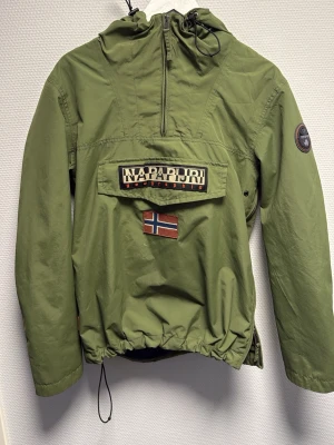 Grön vindjacka från Napapijri - Säljer en grön vindjacka från Napapijri med huva, halv dragkedja och stor ficka framtill med logga och norsk flagga. Jackan har justerbar dragsko nertill och patch på ärmen. Perfekt för dig som gillar stilren och funktionell design. Aldrig använd. Skicka angående frågor!