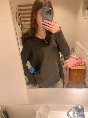 Olivgrön stickad tröja med fuskpälsluva - Snygg olivgrön jacka med stor luva kantad av fuskpäls. Jackan har lång ärm, v-ringning och snörningsdetaljer i sidorna.  Ser brun ut men är oliv mörk grön 🫒💕