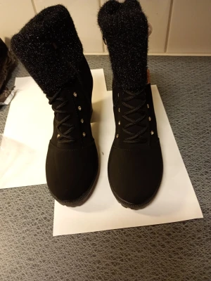 Svarta boots med päls och pärldetalj stl 39  - Säljer ett par svarta boots med grov klack och platt sula. Skorna har glittrig päls runt skaftet, snörning framtill och dekorativa pärlor och strass på sidan. Utsidan är i syntet och insidan är fodrad med mjuk päls. Dragkedja på sidan för enkel påtagning.stl 39 klack 8 cm hög.