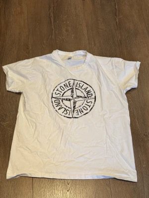 Vit Stone Island t-shirt - Vit t-shirt från Stone Island med stort svart logotryck på bröstet. Klassisk rund halsringning och korta ärmar. Perfekt för en avslappnad och stilren look.