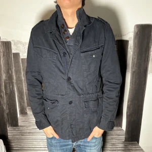 Mörkblå utilityjacka med fickor - Snygg mörkblå utilityjacka med flera stora fickor framtill och axelklaffar. Jackan har krage, knappstängning och långa ärmar. Perfekt för dig som gillar en avslappnad men ändå stilren look.