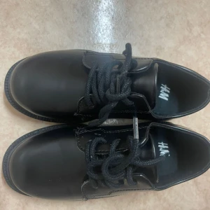 Svarta skor från H&M - Snygga svarta skor från H&M i syntetmaterial. De har en klassisk design med snörning och en rund tå. Perfekta för både vardag och festliga tillfällen. Skorna är i mycket bra skick och redo att användas direkt.