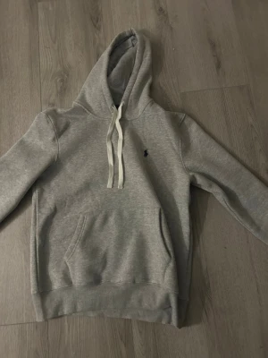 Grå hoodie från Polo Ralph Lauren - Klassisk grå hoodie från Polo Ralph Lauren med vit snörning i huvan och mörkblå logga broderad på bröstet. Hoodien har känguruficka framtill och ribbade muddar vid ärmslut och nederkant. Perfekt för en avslappnad och stilren look. Står storlek S men är väldigt stor så skulle säga medium.