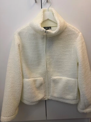 Vit teddyjacka från 157, storlek XS - Mysig vit teddyjacka från 157 i storlek XS. Jackan har hög krage, dragkedja framtill och två stora fickor. Aldrig använd. Nypris 300