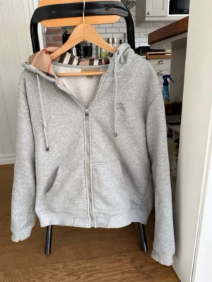 Grå zip hoodie från Burberry - Snygg grå hoodie från Burberry med dragkedja framtill och klassiskt rutigt foder i huvan. Tröjan har två fickor, justerbar huva med snören och ribbade muddar. Perfekt för en avslappnad och stilren look.