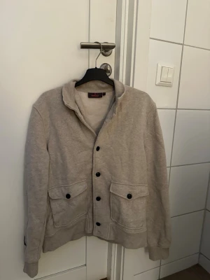 Beige cardigan från Morris - Beige cardigan från Morris med svarta knappar ända upp i halsen. Koftan är i ett superbra skick och är i storlek XS. Det är lite halvsvajigt ljus på bilderna, så man kan få för sig att koftan är ljusgrå men den är beige. Det är bara att hojta till vid frågor!
