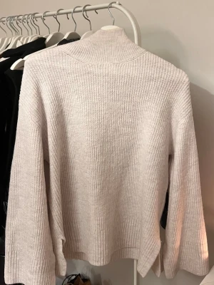 Ljusbeige ribbstickad polotröja - Mysig ljusbeige stickad tröja med hög krage och breda, långa ärmar. Tröjan har ribbstickat mönster och slitsar vid ärmsluten samt nederkanten för en avslappnad look. Perfekt för lager på lager under kyliga dagar.
