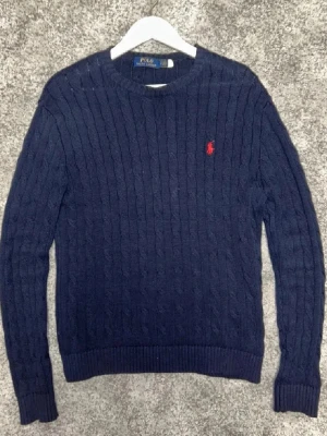 Mörkblå kabelstickad tröja Polo Ralph Lauren🙌🤩 - Mörkblå kabelstickad tröja från Polo Ralph Lauren med rund halsringning och röd broderad logga på bröstet. Tröjan har långa ärmar och ribbade muddar vid ärmslut och nederkant. Perfekt för en klassisk och stilren look.🙌🤩