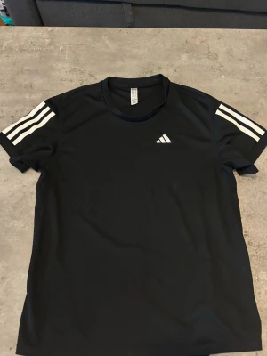 Svart tränings-tshirt från Adidas - Snygg svart tränings-tshirt från Adidas med klassiska vita ränder på axlarna och Adidas-logga på bröstet. Tillverkad i ett lätt och snabbtorkande material som passar perfekt till gymmet eller löprundan.