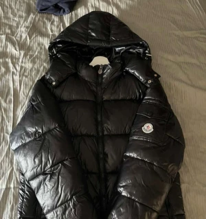 Svart Moncler dunjacka med huva - Snygg svart dunjacka från Moncler med glansig finish och huva. Jackan har dragkedja framtill, två sidofickor med dragkedja och Moncler-logga på ärmen. Perfekt för kalla dagar och ger en riktigt trendig look. Använd en säsong och är i fint skick! 💖