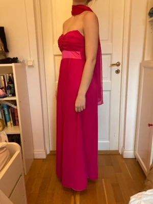 Rosa balklänning - Säljer en långklänning i ceriserosa nyans med draperad överdel och bred satinliknande midjedetalj. Axelbanden är avtagbara. Klänningen har ett flytande material som ger en snygg siluett. Schalen ingår och går att ha på massa olika sätt! Priser är diskuterbart