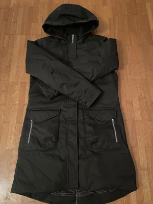 Grön parkasjacka från Elvine - Snygg grön parkasjacka från Elvine med huva och dragkedja framtill. Jackan har stora fickor med dragkedjor och en längre passform. Insidan är fodrad för extra värme och komfort, perfekt för kyliga dagar.