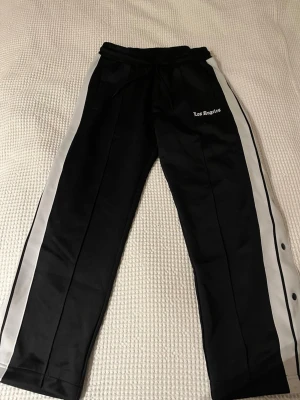 Svarta mjukisbyxor med vita ränder - Snygga svarta mjukisbyxor med vita ränder längs sidorna och trycket 'Los Angeles' på ena benet. Byxorna har resår i midjan med snörning och tryckknappar längs sidan för extra stilpoäng. Perfekta för en chill och sportig look.