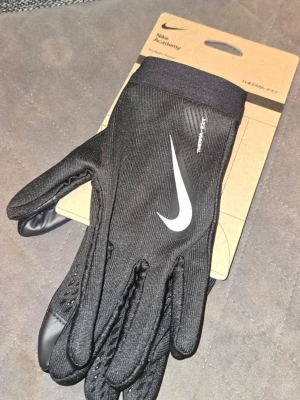 Svarta Nike Academy handskar - Snygga svarta handskar från Nike Academy med vit logga på ovansidan. Tillverkade i ett stretchigt material med Therm-FIT-teknologi som håller händerna varma. Perfekta för träning eller vardag när det är kallt ute. Priset är satt. Helt nya handskar. Hör av er vid frågor.