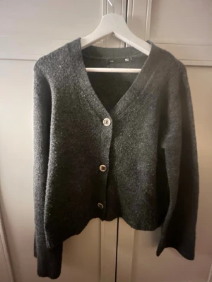 Mörkgrå kofta från 157 med knappar - Mysig mörkgrå kofta från 157 med tre stora knappar framtill och djup v-ringning. Koftan har lång ärm och är tillverkad i ett mjukt, stickat material som passar perfekt för lager-på-lager-stil.