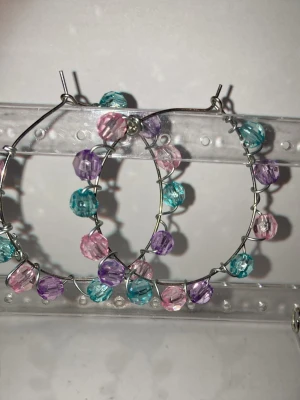 Hoops med färgglada pärlor - Handgjorda örhängen som görs av mig i silverfärgad metall med plast pärlor i lila, rosa och turkos. Pärlorna är jämnt fördelade runt hela örhänget och ger en lekfull och trendig look. Perfekta för att addera färg till din outfit. Frakten är ca22-100 kr beroende på vikten. Kontakta mig om du vill köpa eller har frågor.