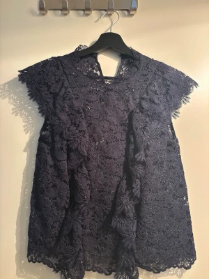 Mörklila spetsblus med volanger - Säljer en mörklila blus i vacker spets med blommönster och dekorativa volanger framtill. Blusen har rund halsringning och korta ärmar. Perfekt för dig som gillar romantisk och trendig stil.