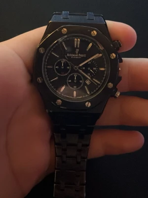 Svart Audemars Piguet Royal Oak klocka - Snygg svart Audemars Piguet Royal Oak herrklocka med länkarmband i metall. Klockan har kronograf, datumvisning och tydliga indexmarkeringar. Boetten är åttakantig med synliga skruvar och urtavlan är svart med silverdetaljer.