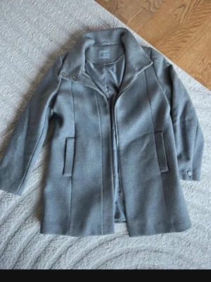 Grå kappa från Lindex, storlek M - Snygg grå kappa från Lindex i storlek M. Rak modell med hög krage som kan knäppas med tryckknappar, dolda fickor och slät insida. Perfekt för dig som gillar stilrena och klassiska plagg. Materialet känns mjukt och har en ullig look.