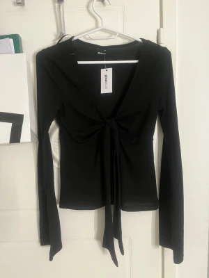 Svart knytblus från Gina Tricot - långärmad topp från Gina Tricot i tunn, mjuk polyester. Modellen har djup v-ringning och knyt framtill som ger en snygg siluett. I stolek xs