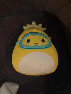 Gul ananas Squishmallow  - Supergulligt Squishmallow mjukdjur format som en ananas med blåa simglasögon och ett leende ansikte. Tillverkad i mjuk plysch och har rutmönster i orange. Perfekt som prydnad eller kramkompis!